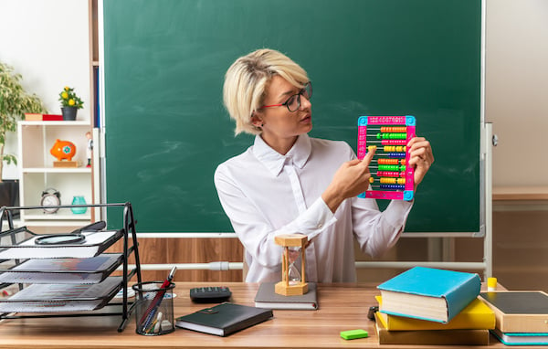 Young Blonde Female Teacher Wearing Glasses Sitting At Desk With School Tools In Classroom Showing Abacus Looking At It Pointing Finger On It