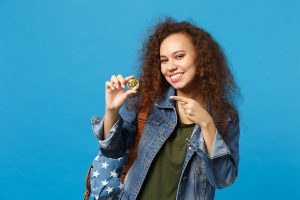 Young African American Girl Teen Student In Denim Clothes, Backpack Hold Bitcoin Isolated On Blue Background Studio Portrait. Education In High School University College Concept. Mock Up Copy Space.
