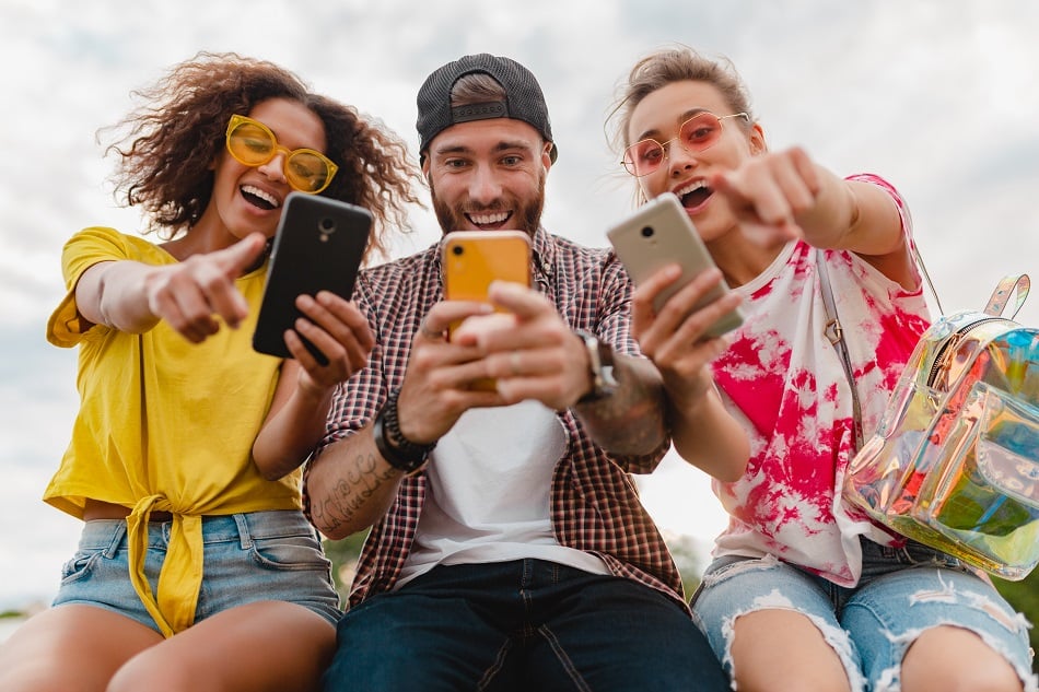 Happy Young Company Of Smiling Friends Sitting Park Using Smartphones, Man And Women Having Fun Together, Colorful Summer Hipster Fashion Style, Communication Wireless Connecting Devices